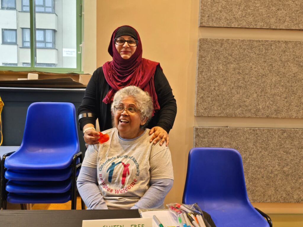 Two women smiling, one sitting on a chair and the other standing behind her with her hands on the first woman's shoulders
