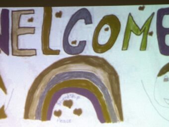 Sign hand-drawn by children which says Welcome and includes drawings of a rainbow, two faces and a star.