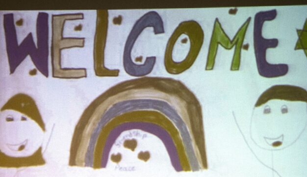 Sign hand-drawn by children which says Welcome and includes drawings of a rainbow, two faces and a star.