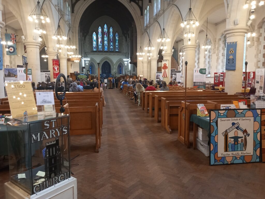 Image at the back of St Mary's Church, with the Swansea Schools of Sanctuary logo mural in the foreground, with students and teachers sitting in the background, facing the stage area. 